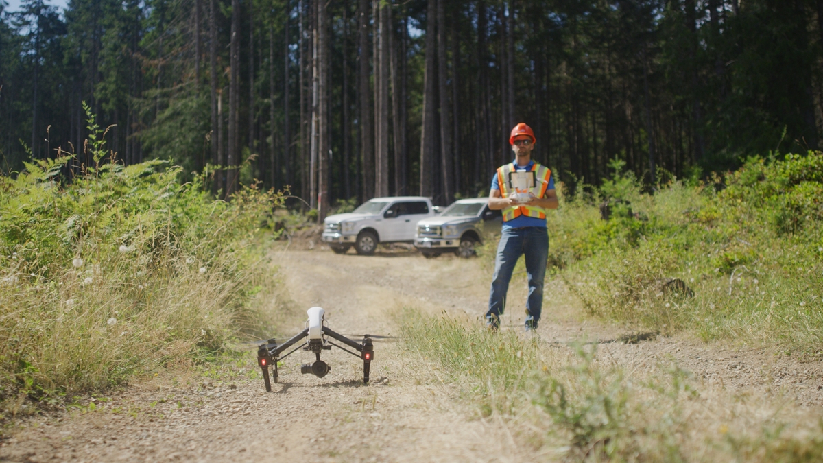 Outdoor sunny image of forestry survey worker in PPE standing on dirt road with forested area in background, aerial drone in take-off position, and controller in hand