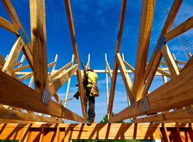 Light-frame construction beams and roof trusses shown being installed on low-rise residential structure by construction worker with nail gun and fall arrest harness