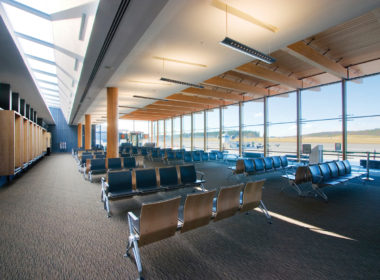 Interior daytime view of low rise Prince George Airport Expansion showing blue chairs, sculpted ceiling and structural post + beam timbers supporting Glue-laminated timber (Glulam)
